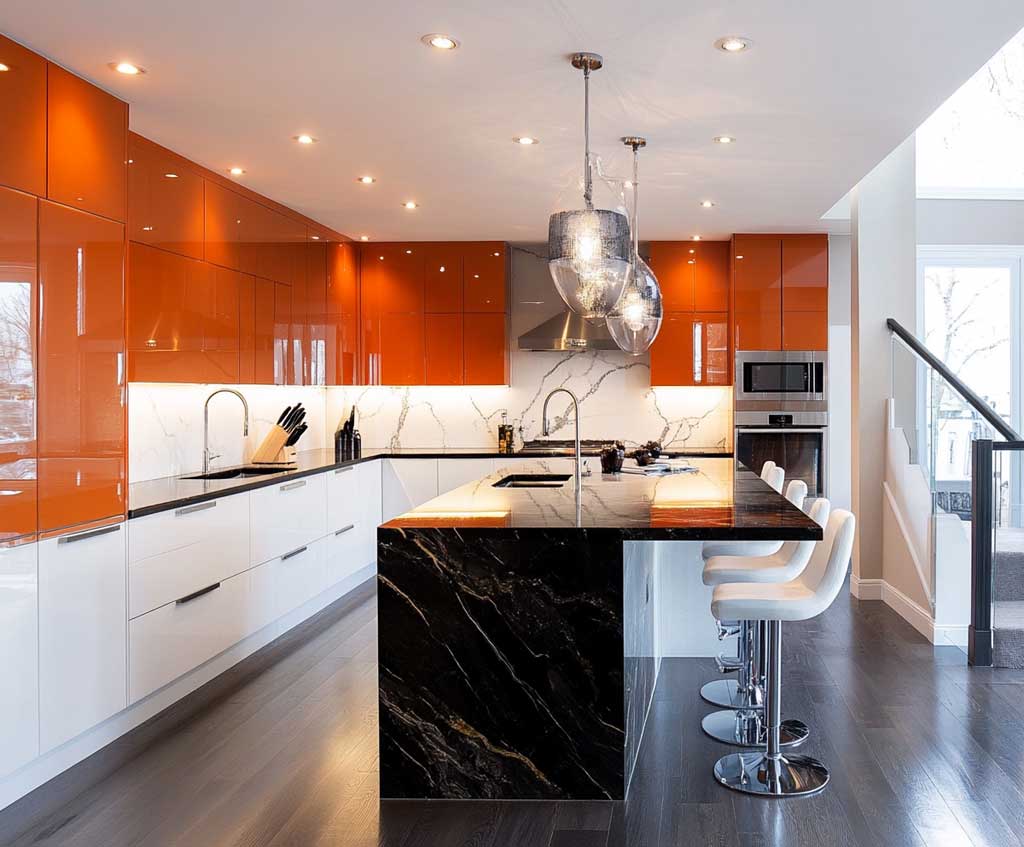 A modern L-shaped kitchen featuring an elegant black marble island paired with glossy orange upper cabinets. White lower cabinets balance the vibrant color scheme. Warm ambient lighting illuminates the kitchen, with dark wood flooring adding texture and richness to the overall design.
