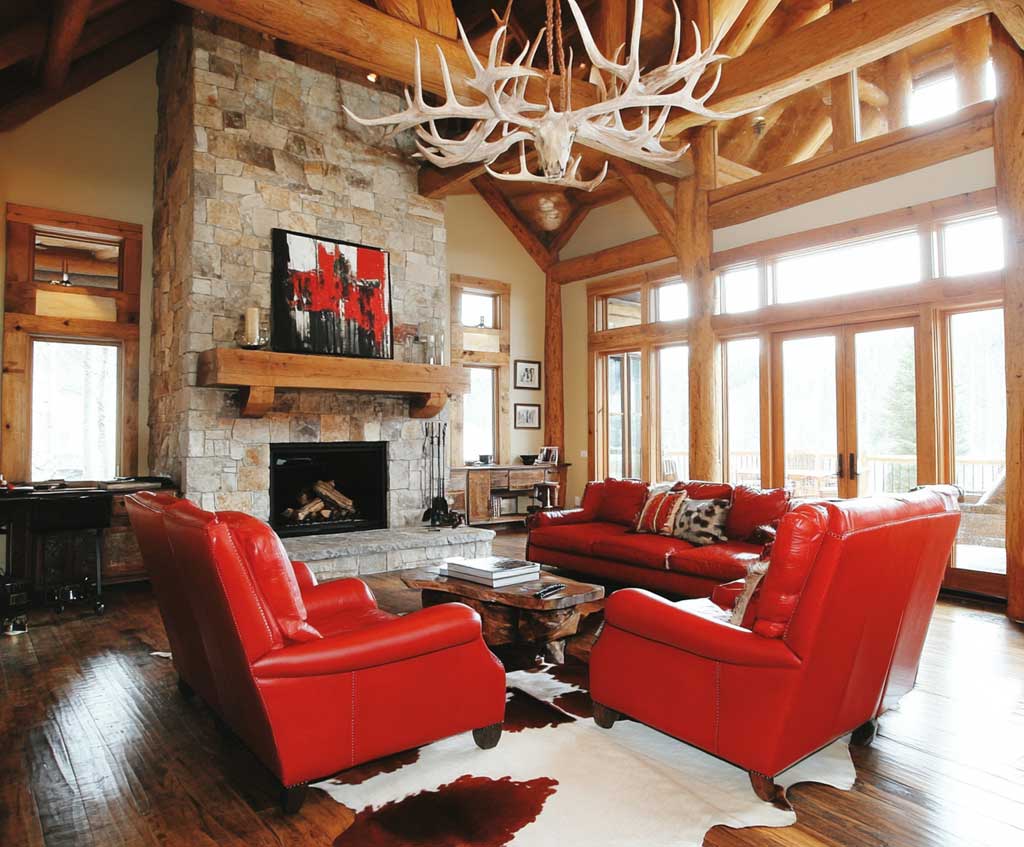 A spacious living room featuring rustic wooden beams, a pair of bold red leather armchairs, a vintage cowhide rug, a stone fireplace, and antler chandeliers. The armchairs stand out against the neutral tones of the wood and stone, adding a vibrant, fiery touch to the cozy western atmosphere.