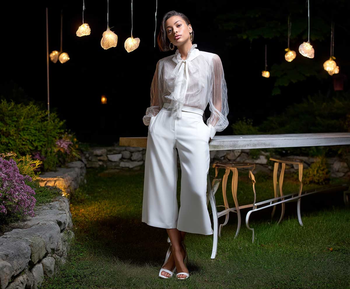 Chic woman at a garden cocktail party, wearing a structured organza sheer white blouse tucked into belted white culotte pants, dramatic lighting, modern garden with floral lights, elegant heels, full-length editorial photograph