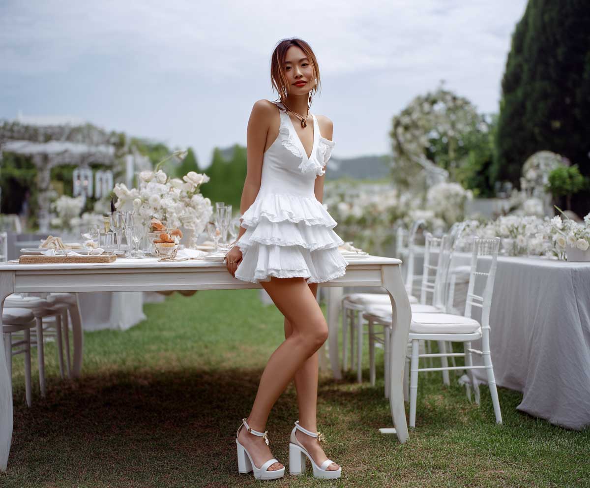 Fashionable woman at an elegant outdoor brunch party, wearing a white tiered ruffle mini dress and chunky white block heels, sun-kissed setting, floral table setups, soft breeze, cinematic full-length fashion photograph