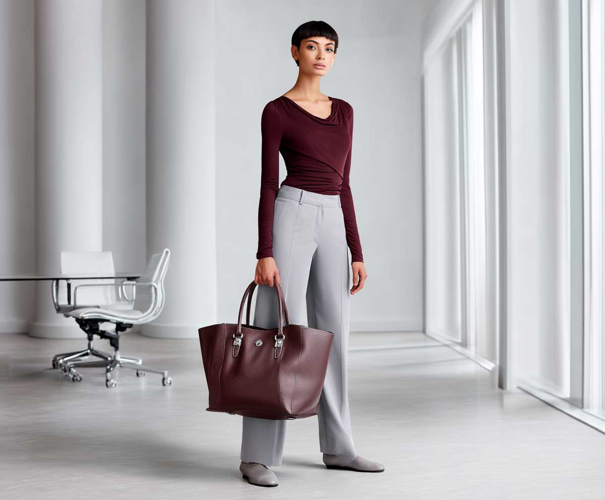 fashionable female model wearing burgundy wrap top with light gray straight leg trousers, carrying a structured leather tote, soft modern office setting, clean lighting, relaxed yet polished style