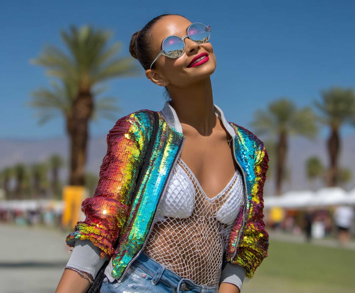 Smiling model in rainbow sequin bomber jacket, white fishnet layers, denim shorts, bold makeup, reflective sunglasses, palm trees in the distance, vibrant music festival style