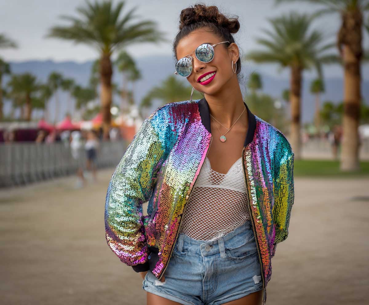 Smiling model in rainbow sequin bomber jacket, white fishnet layers, denim shorts, bold makeup, reflective sunglasses, palm trees in the distance, vibrant music festival style