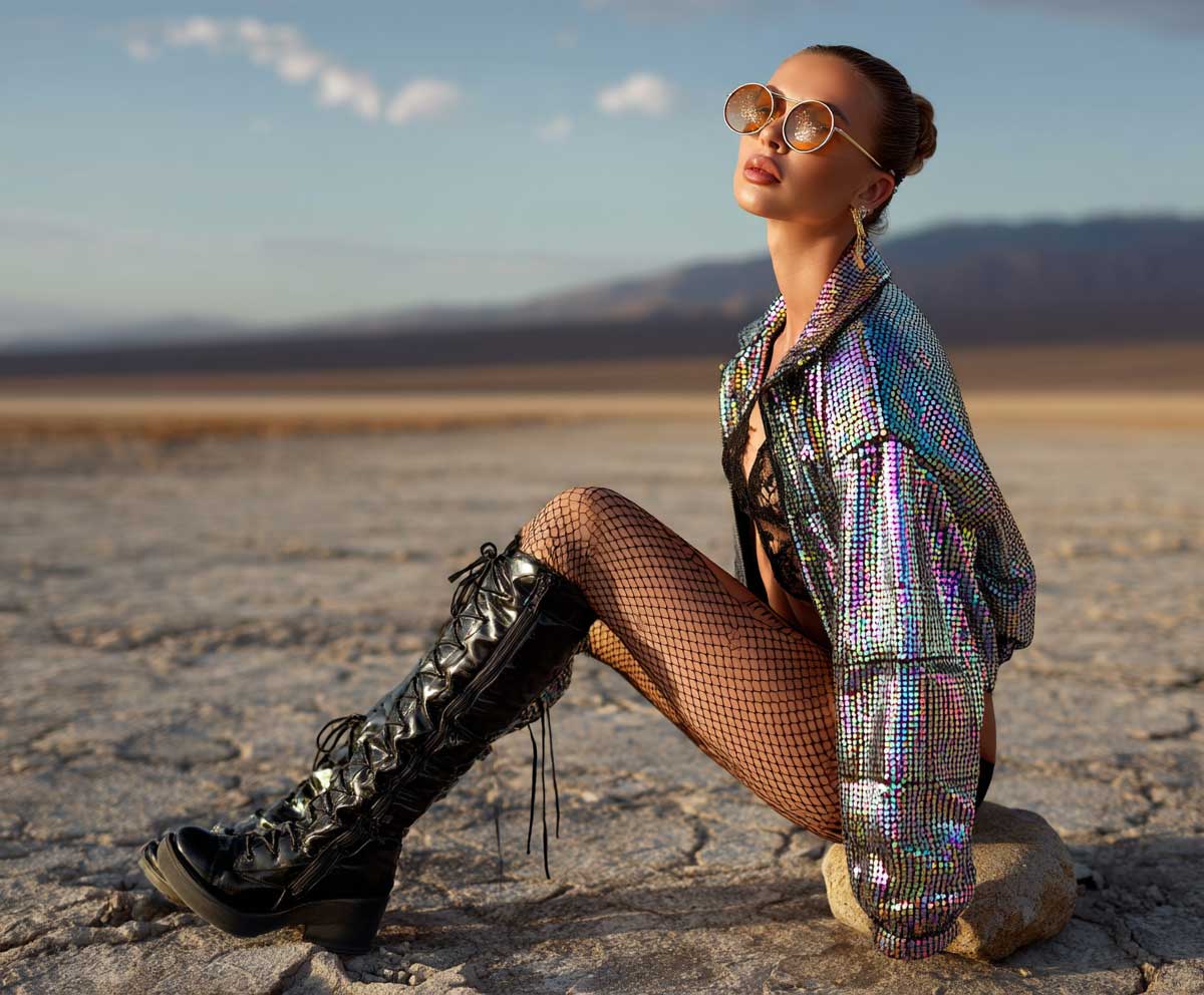 Festival model in holographic sequin jacket, black fishnet tights, black bodysuit, high boots, desert background, edgy glam look, golden hour lighting, full-body Coachella fashion photo