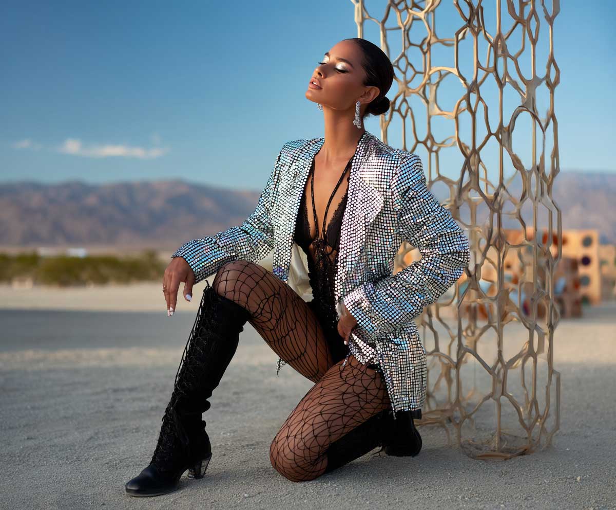 Festival model in holographic sequin jacket, black fishnet tights, black bodysuit, high boots, desert background, edgy glam look, golden hour lighting, full-body Coachella fashion photo