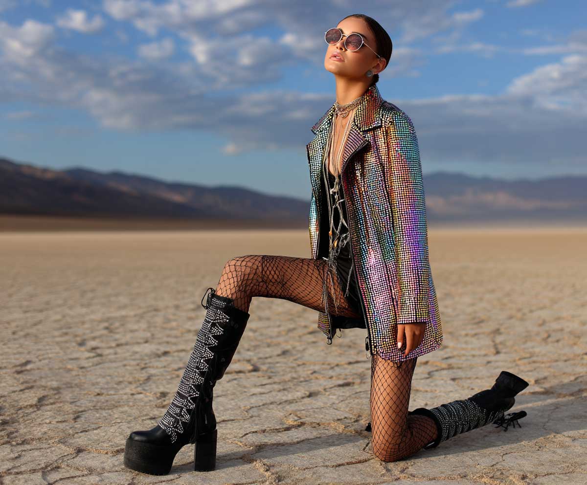 Festival model in holographic sequin jacket, black fishnet tights, black bodysuit, high boots, desert background, edgy glam look, golden hour lighting, full-body Coachella fashion photo