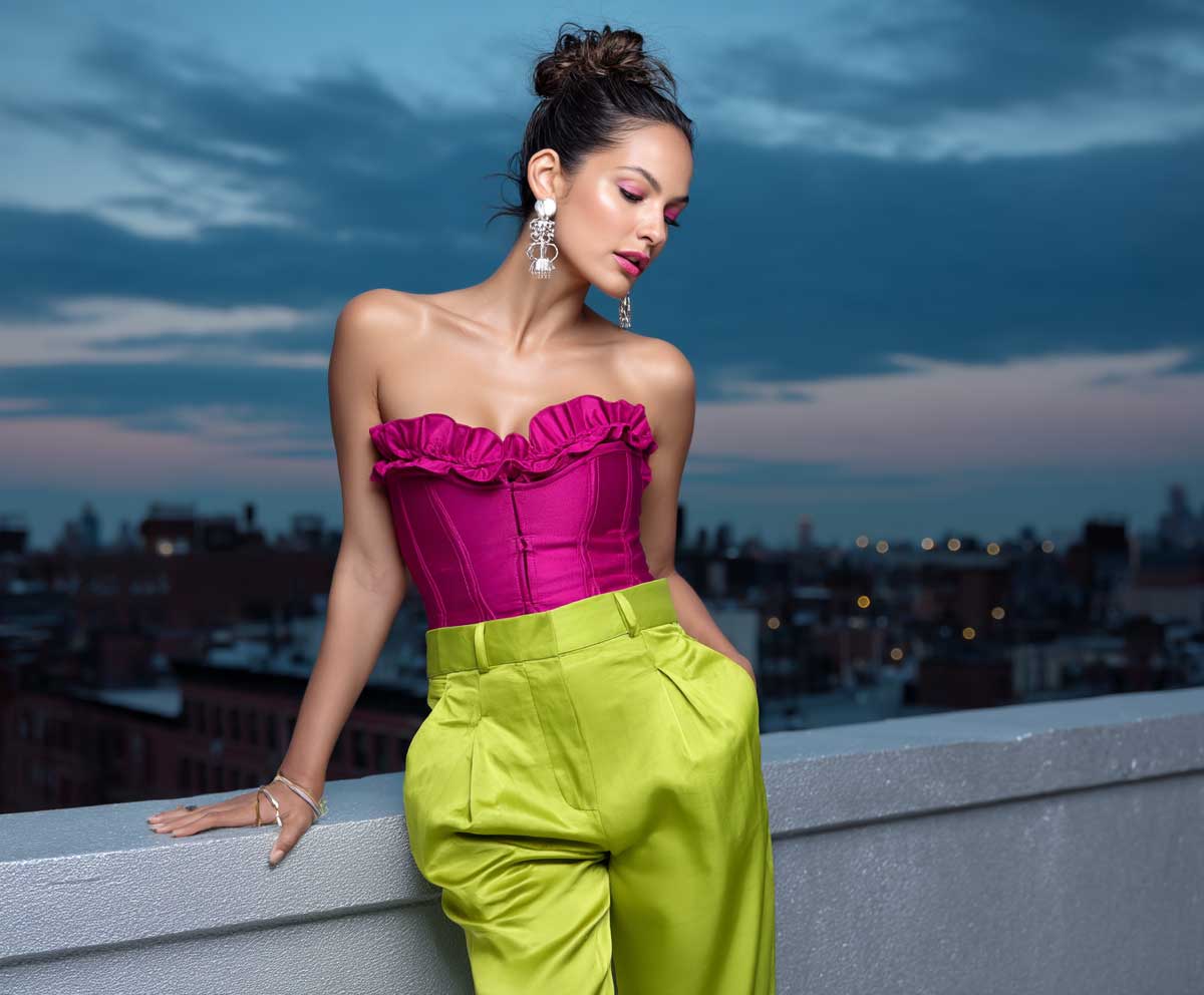stylish woman posing in lime green satin trousers and a structured fuchsia corset top, rooftop city background, twilight sky, glowing makeup, chic evening vibe, fashion editorial