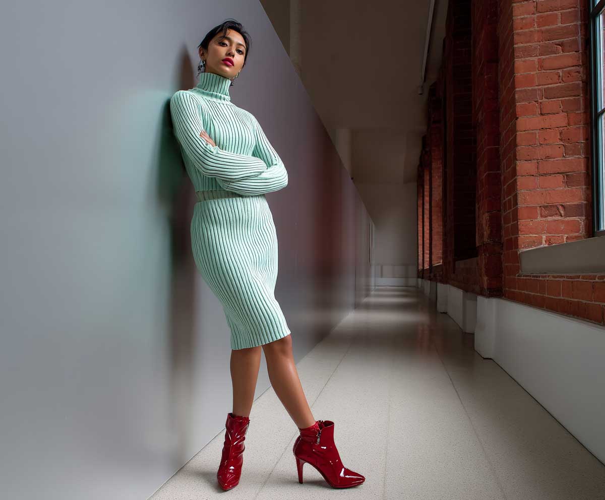 woman in a form-fitting mint green knit dress paired with glossy ruby red ankle boots, art gallery backdrop, indoor moody lighting, confident pose, cozy yet bold fashion vibe, editorial style