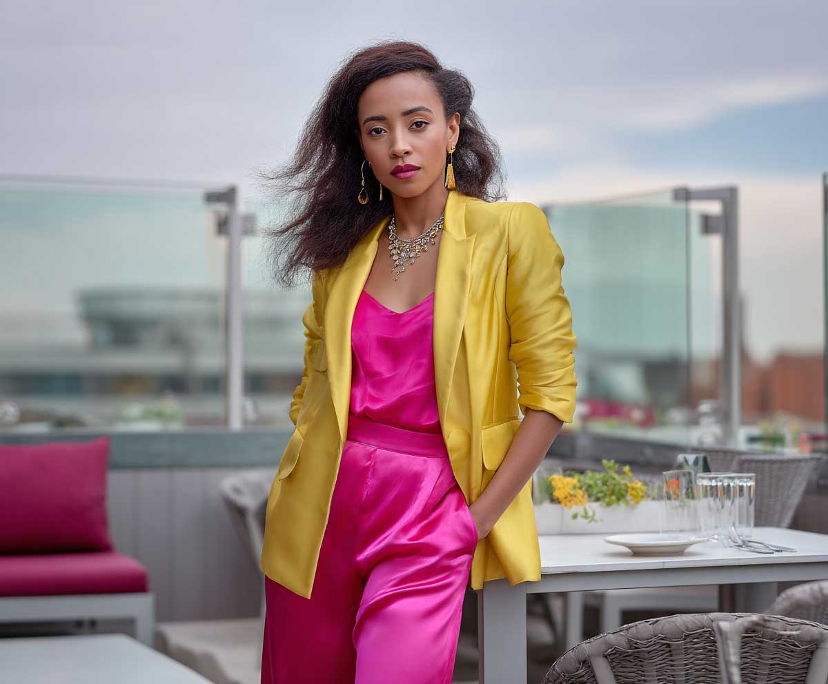 confident woman in a canary yellow blazer layered over a hot pink satin jumpsuit, rooftop restaurant setting, sunset sky, soft wind in hair, chic and vibrant style, natural makeup look