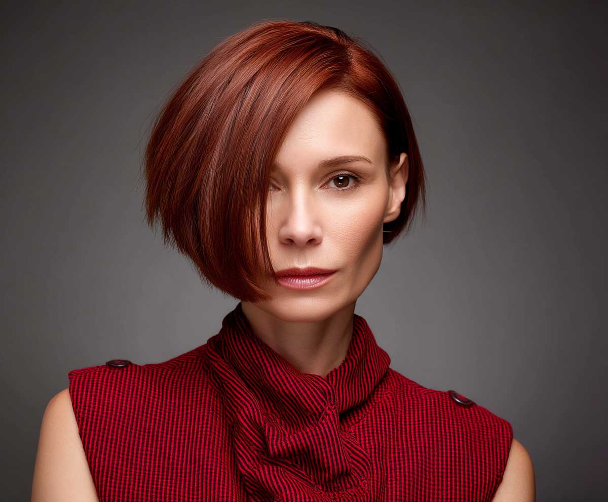 Elegant woman with a square face, short straight asymmetrical crop in auburn red, contemporary studio photo, clean lighting, strong posture