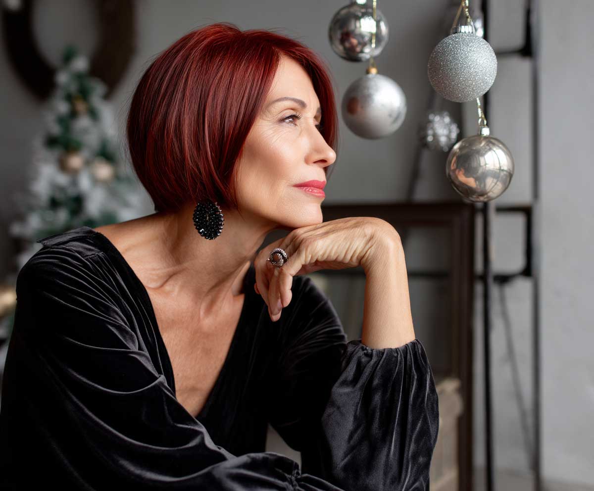 stylish woman over 50 with sleek cherry red long bob haircut, holiday party decorations background, elegant black velvet dress, soft evening light