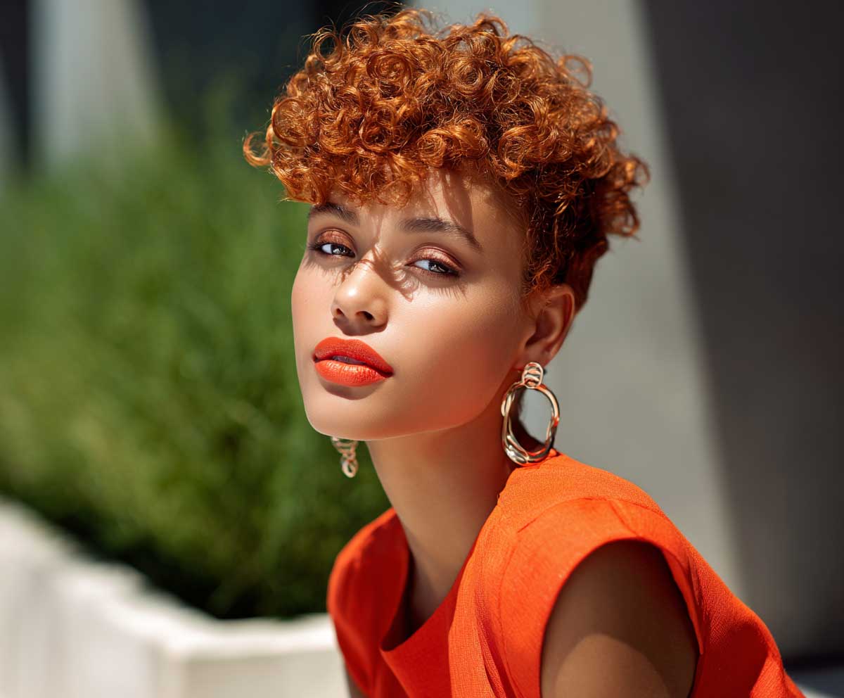 editorial-style portrait of a young woman with a bright coral curly pixie cut, voluminous curls at the crown, glowing skin, coral lips, sunlit outdoor setting, modern hairstyle focus
