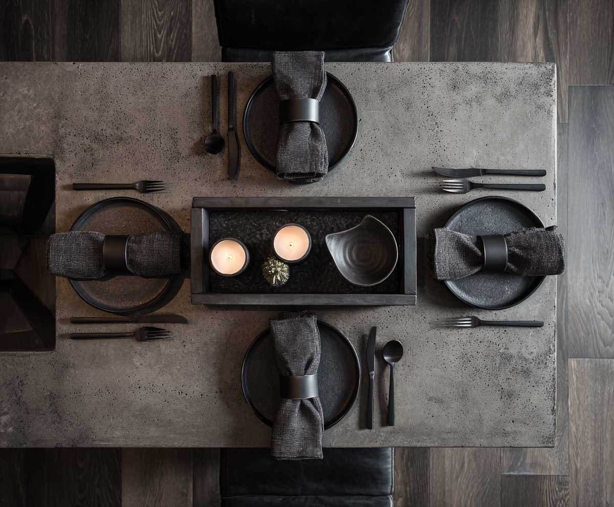 dining table setting featuring concrete table top, matte black ceramic plates, iron napkin rings, minimalist industrial style, soft overhead light, dramatic contrast, dark wood floor, high detail