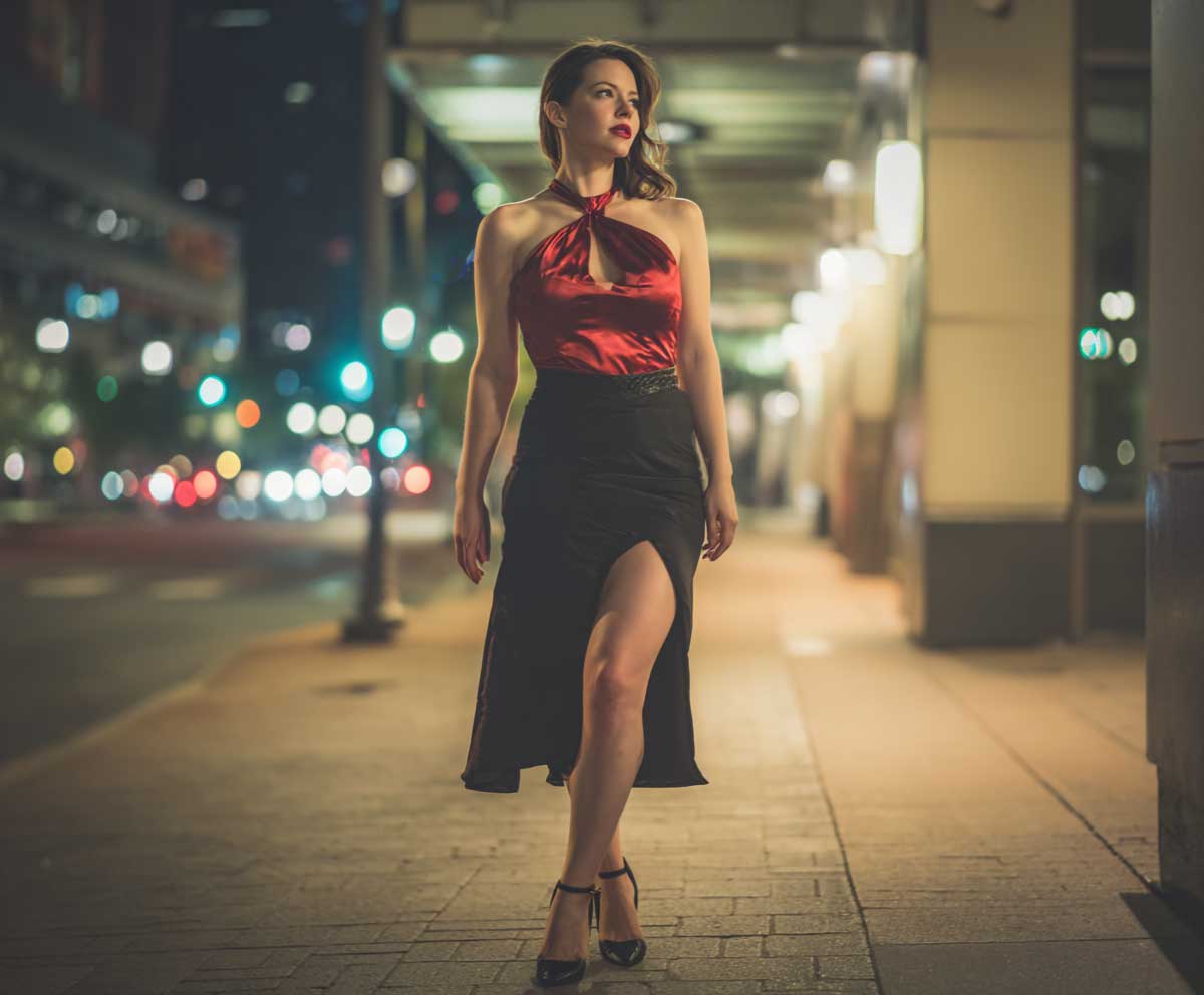 full-body shot of a confident woman wearing a red satin halter top and a sleek black skirt with a thigh-high slit, moody evening lighting, glossy heels, nighttime city street backdrop, soft shadows, minimal accessories