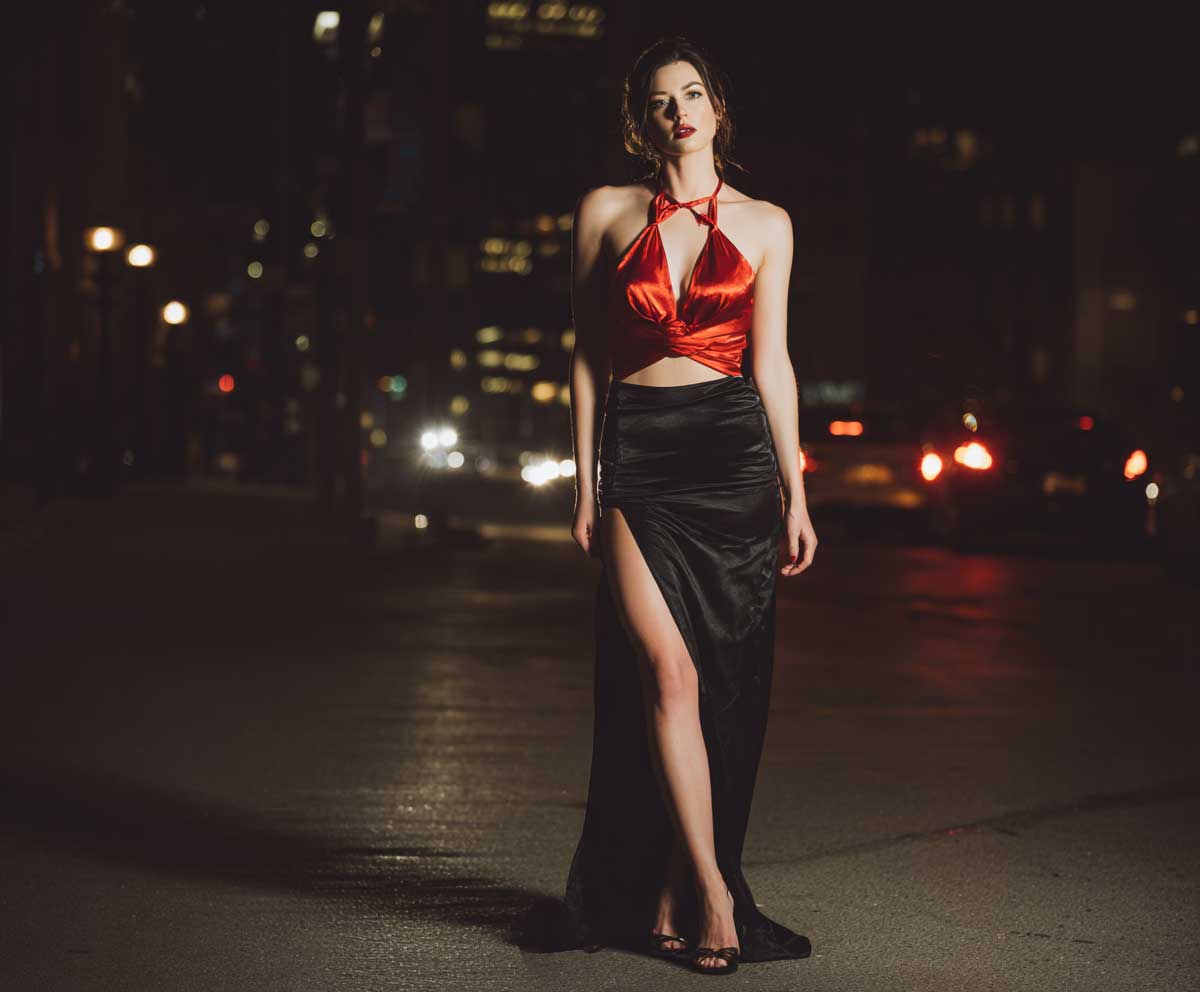 full-body shot of a confident woman wearing a red satin halter top and a sleek black skirt with a thigh-high slit, moody evening lighting, glossy heels, nighttime city street backdrop, soft shadows, minimal accessories