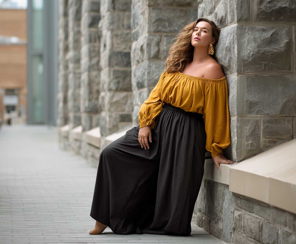 plus size woman in black palazzo pants and a mustard off shoulder top, leaning against a stone wall, urban backdrop, soft wind in hair, modern full body fashion photo, moody lighting