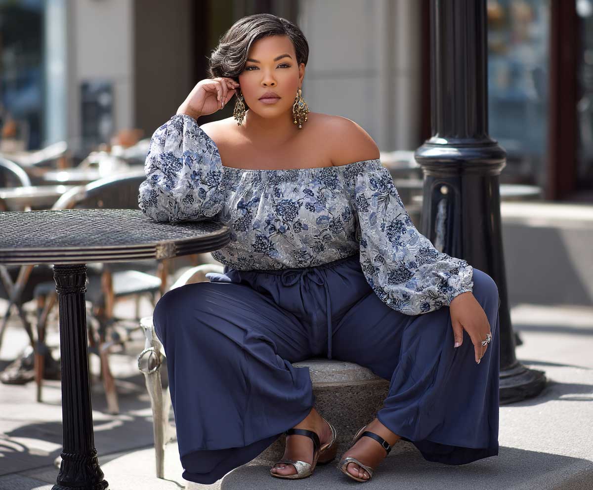 plus size woman wearing navy palazzo pants and a floral off shoulder blouse, sitting on a cafe terrace, late afternoon sunlight, soft shadows, elegant casual style, full body view