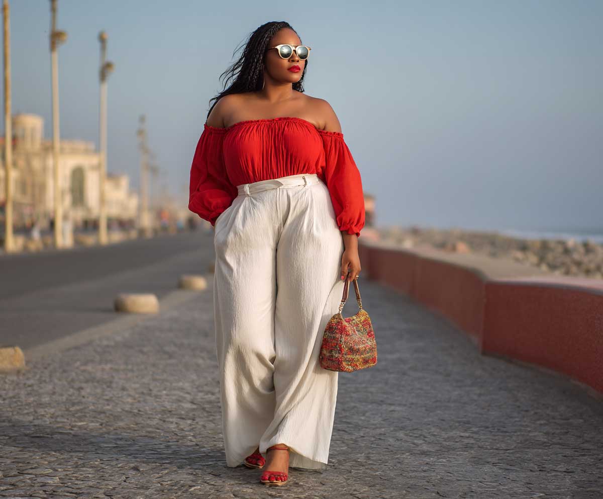 plus size woman wearing white palazzo pants and a red off shoulder top, standing on a coastal promenade, full body outfit, golden hour, confident pose, vibrant summer fashion style