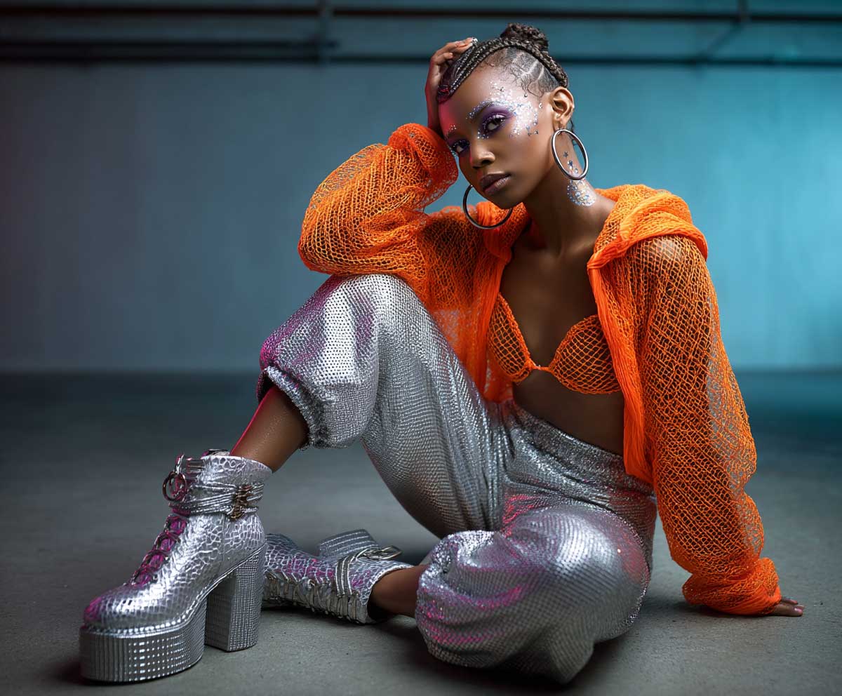 fashion photoshoot of a model at a neon-lit rave, wearing an orange neon mesh hoodie layered over a bandeau, wide-leg silver pants with reflective texture, platform boots, UV body paint, sharp lighting contrast, cyber rave vibe