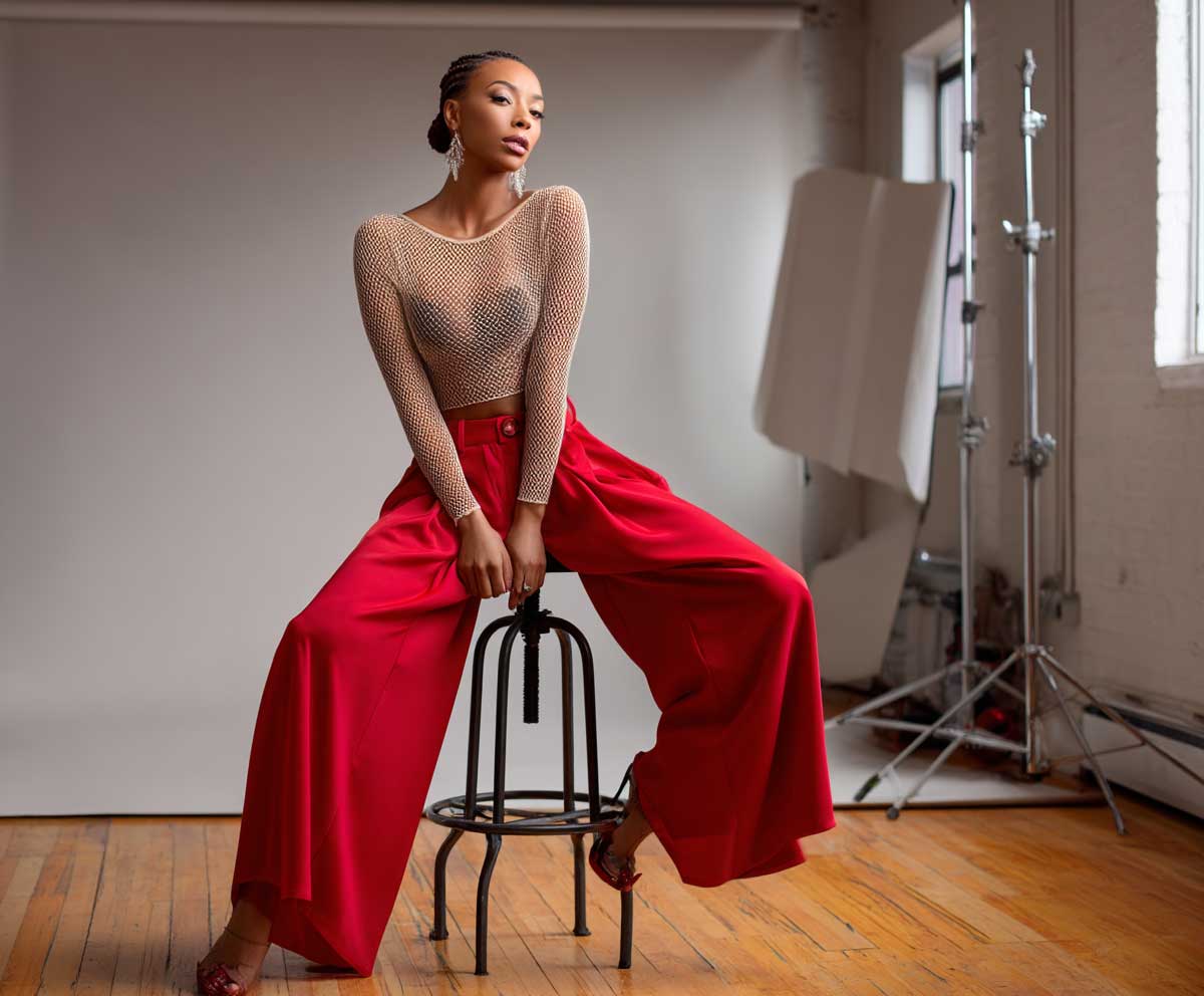 woman wearing soft beige mesh bodysuit and high waist red wide leg trousers, fashion studio setup, bold color styling, relaxed confident posture, warm lighting, full body look