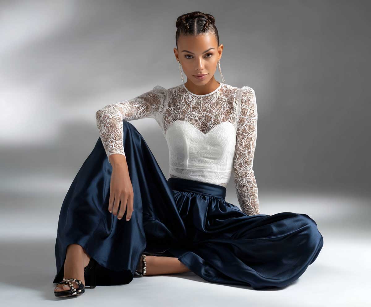 model in white bodysuit with sheer panel details and navy satin wide leg trousers, polished studio lighting, minimal backdrop, soft posture, fashion-forward styling, full length view