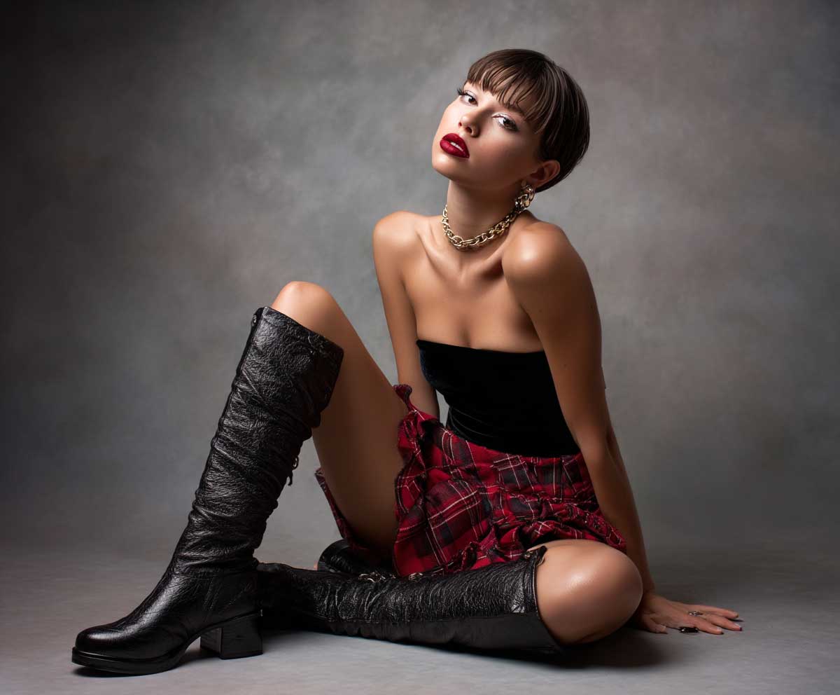 nighttime studio fashion photo of a model in a black velvet tube top and red plaid micro skirt, tall boots, dark red lipstick, sharp lighting, smoky background, edgy expression, high fashion mood