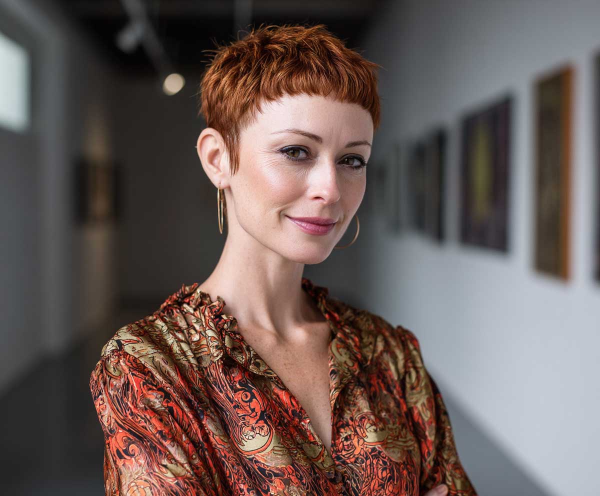 portrait of a creative woman with a warm auburn short pixie cut, edgy abstract micro fringe, standing in a contemporary art gallery, wearing a printed silk blouse, warm tones, Leica camera style, editorial look