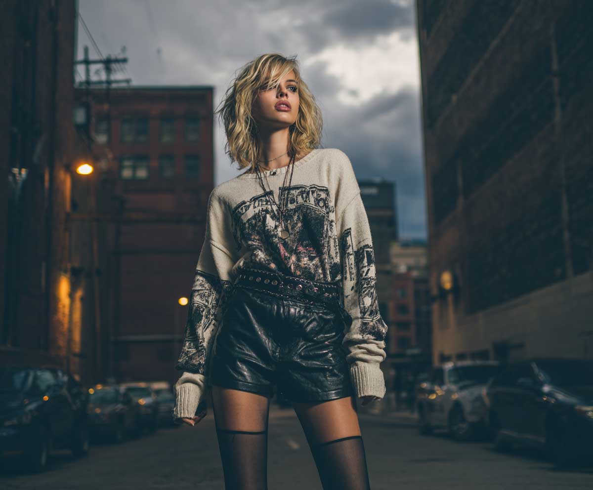 fashionable woman in oversized band graphic sweater with black leather shorts and sheer tights, gritty street fashion aesthetic, evening city background, moody lighting, edgy outfit, urban rock look, full body stylish pose