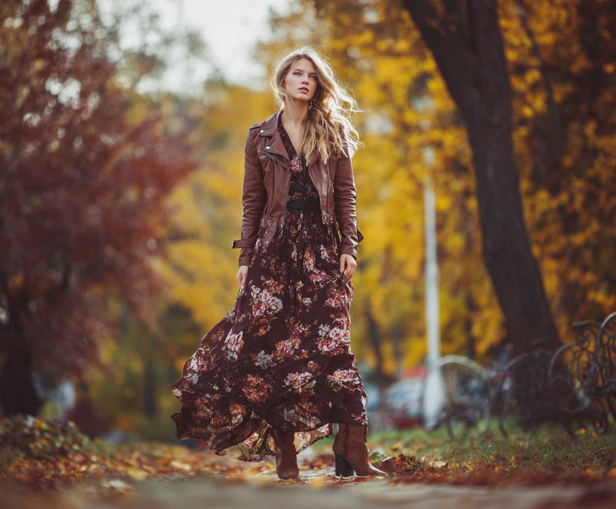 fashion-forward woman in brown leather jacket with dark floral maxi dress, ankle boots, autumn park scene, soft focus background, layered outfit look