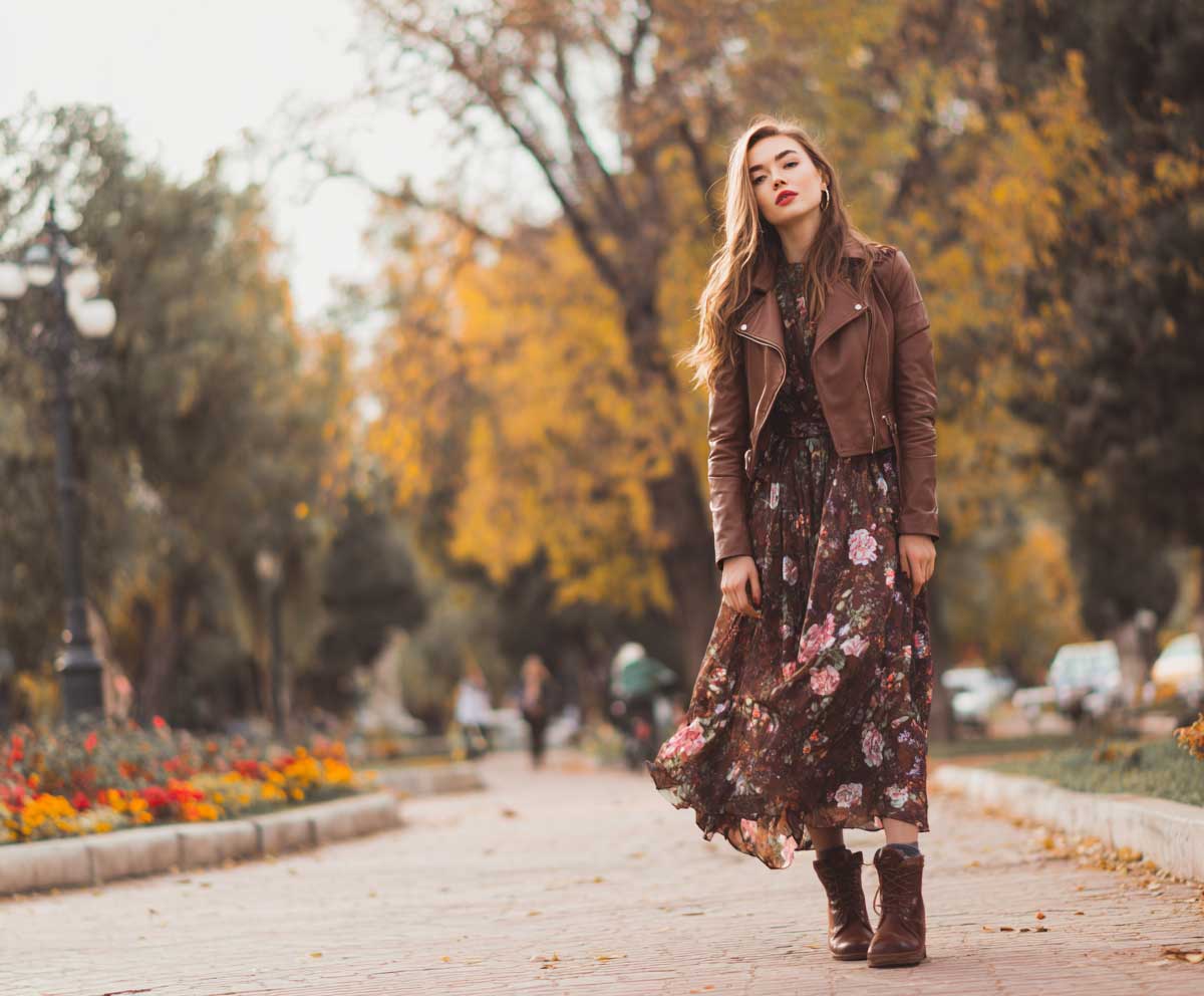 fashion-forward woman in brown leather jacket with dark floral maxi dress, ankle boots, autumn park scene, soft focus background, layered outfit look