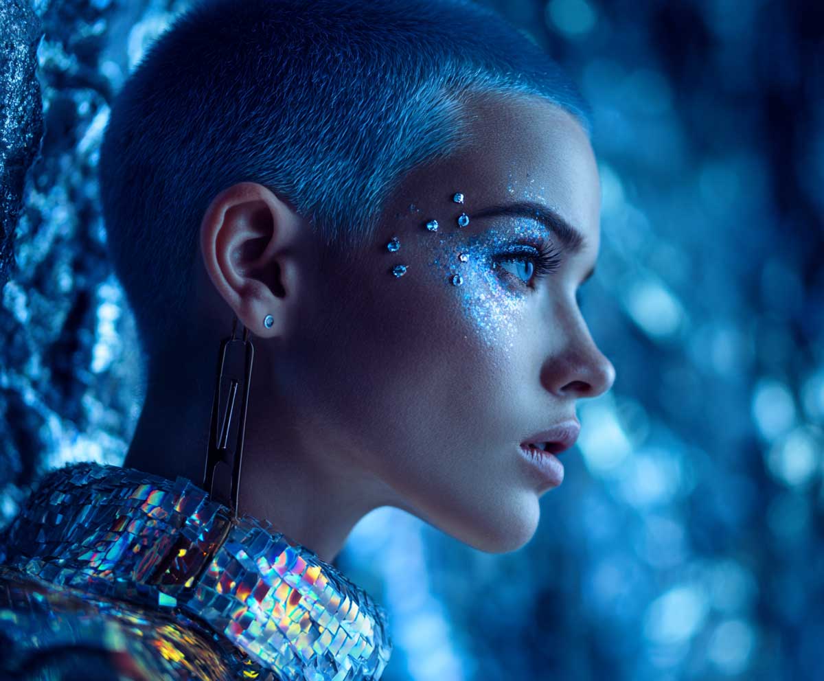 buzz cut dyed in electric blue, futuristic makeup, holographic costume, silver face gems, outer space background, glowing effect, modern short hair look, neon tones, fantasy setting