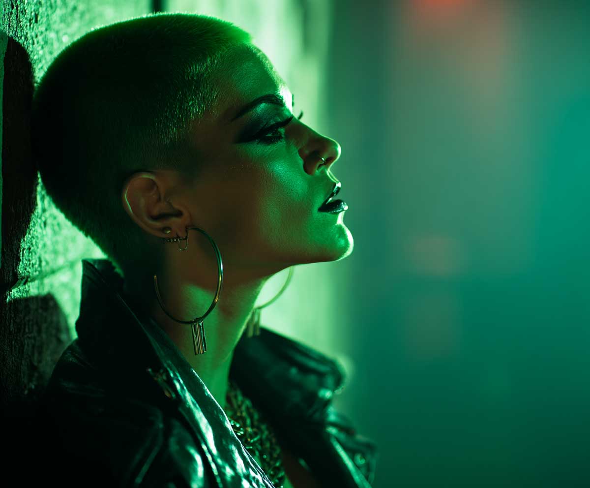 short side-shaved hair dyed neon green, villain-inspired costume, smoky eye makeup, dark leather jacket, eerie green lighting, city alley background, high contrast bold style, Halloween theme