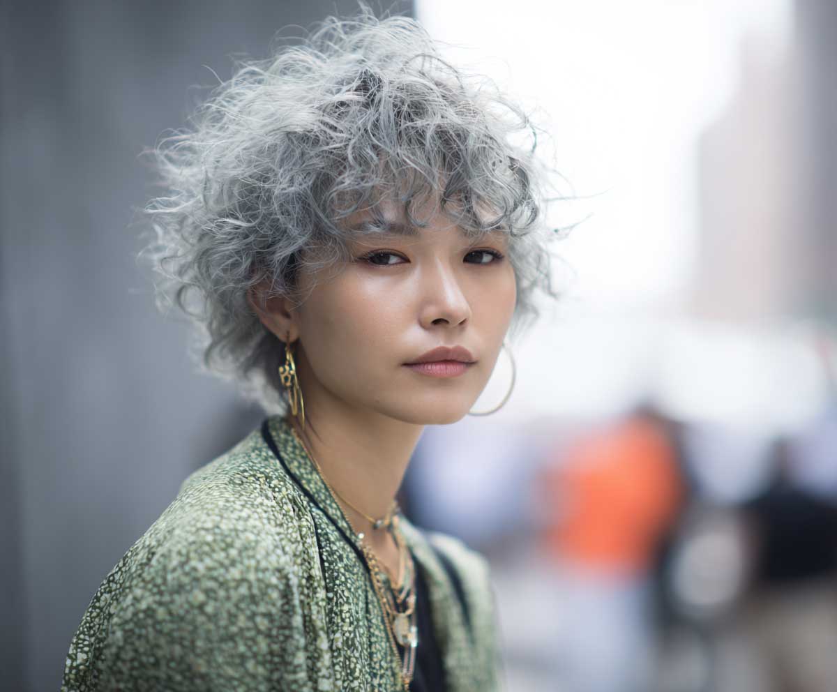 portrait photo of a woman with a layered Korean short hair perm in muted ash green, feathered curls, light texture, wispy bangs, soft natural lighting, street fashion vibe