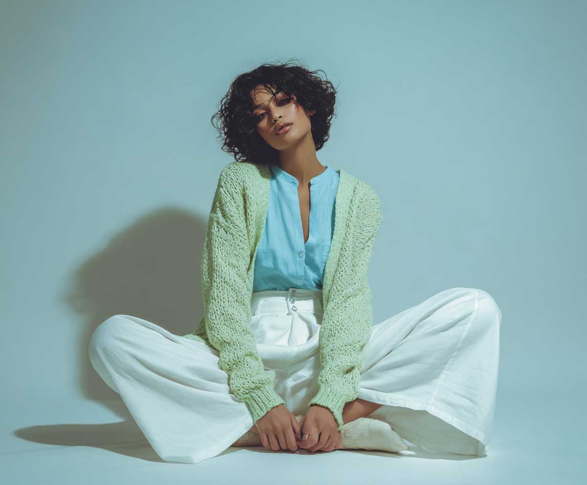 Studio fashion photo of model in mint green cardigan, sky blue blouse, and white linen trousers, minimalist pastel background, relaxed pose, spring vibes