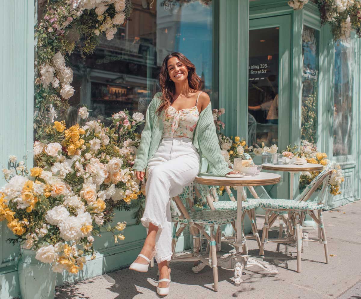 Full-body photo of smiling woman in pastel floral top, mint green cardigan, and white linen pants, seated at a sidewalk café surrounded by spring flowers, fresh daylight, lifestyle editorial