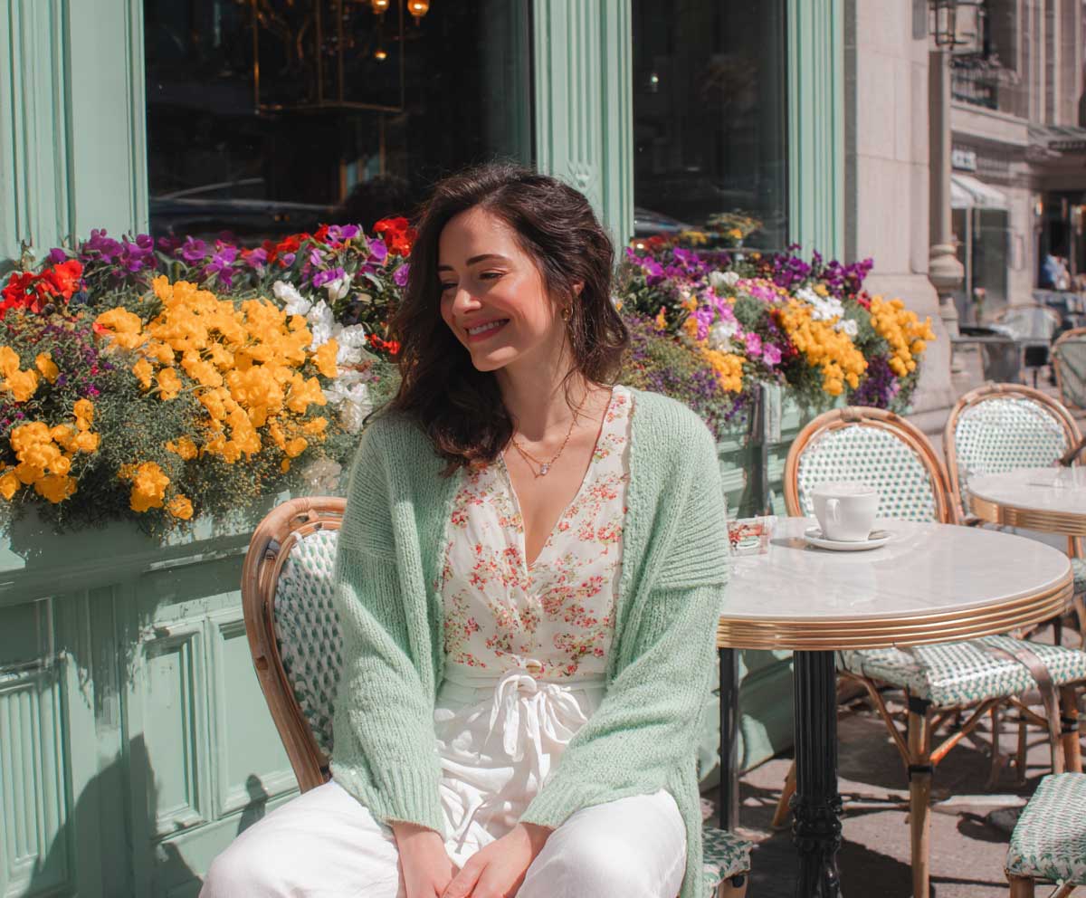 Full-body photo of smiling woman in pastel floral top, mint green cardigan, and white linen pants, seated at a sidewalk café surrounded by spring flowers, fresh daylight, lifestyle editorial
