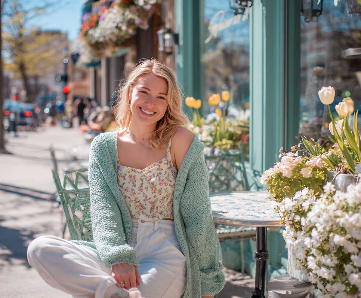Full-body photo of smiling woman in pastel floral top, mint green cardigan, and white linen pants, seated at a sidewalk café surrounded by spring flowers, fresh daylight, lifestyle editorial